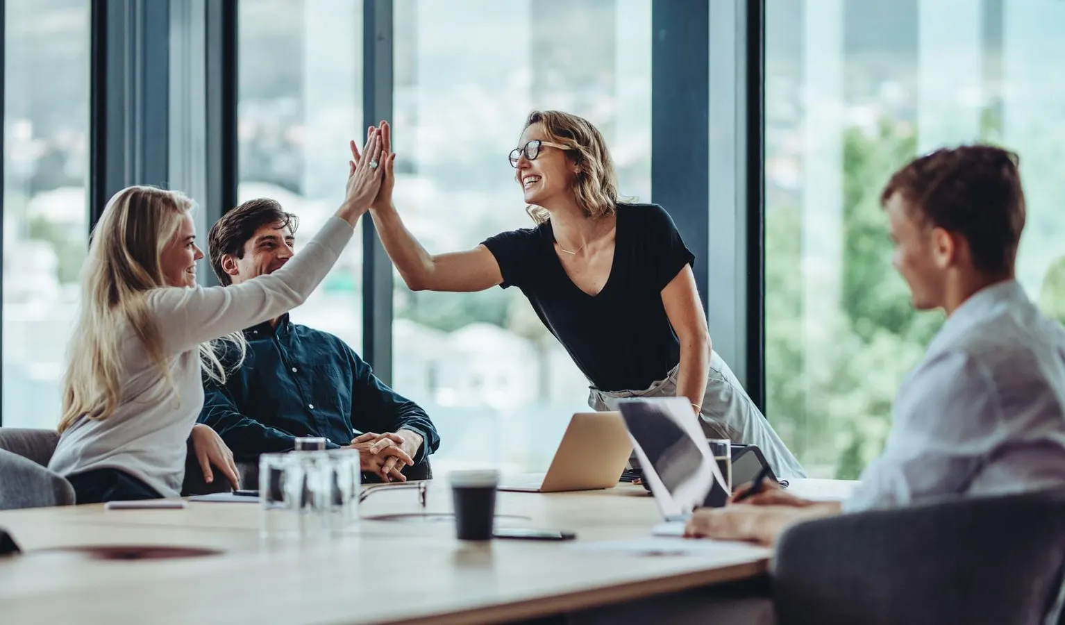 Four colleagues smiling around a meeting table as two teammates share a high five.